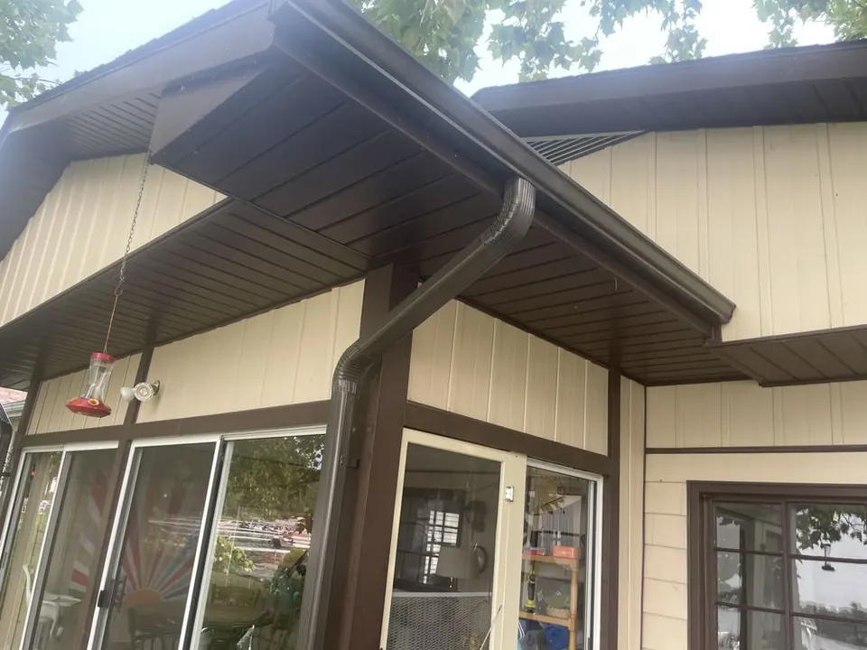 Dark brown gutter along a porch roof with a downspout angled to a corner post